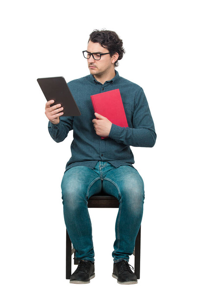 Ciekawy uczeń lub nauczyciel, siedzący na krześle za pomocą tabletu PC, trzymając czerwoną książkę. Wybór pomiędzy tradycyjnym podręcznikiem a nowoczesnym ebookiem, odizolowanym na biało. Koncepcja dylematu edukacji. - Zdjęcie, obraz