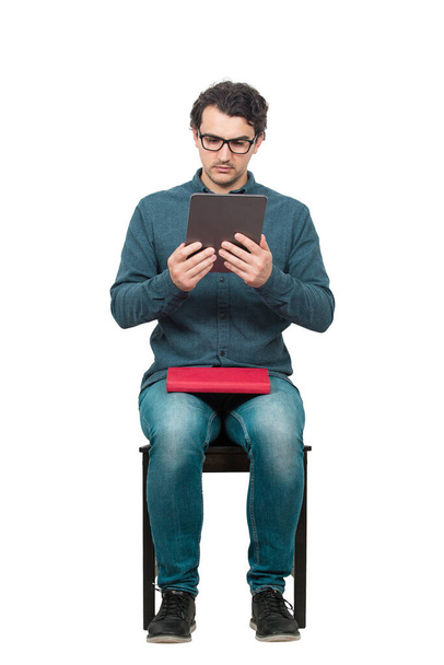 Curious student man seated on a chair using a PC tablet while holds a red book on his knees. Choose modern computer education instead traditional textbook, isolated on white. Study and work from home. - Photo, image