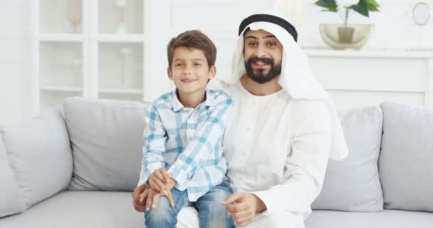 Portrait of young handsome muslim father in kandura sitting on couch in living room with cute small son on laps, talking, waving with hands and smiling to camera. Dad with little boy on sofa at home. - Filmagem, Vídeo