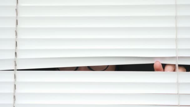 Mujer en gafas mirando furtivamente a través de las persianas blancas
 - Metraje, vídeo