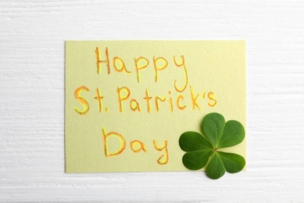 Clover leaf and note with text HAPPY ST. PATRICK'S DAY on white wooden table, flat lay - Foto, Imagem