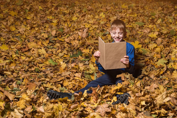 Menino sentado em folhas caídas lê livro e ri. Criança com livro na floresta de outono
. - Foto, Imagem
