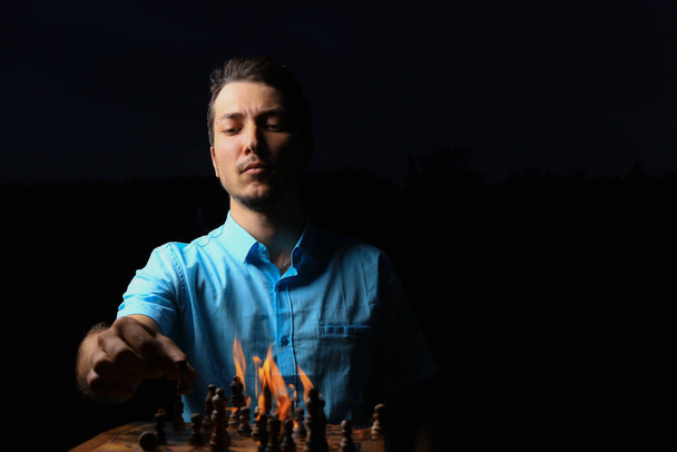 Hombre joven blanco caucásico o chico adulto y un tablero de ajedrez en llamas. Llave baja oscura, fondo negro, enfoque selectivo. - Foto, Imagen