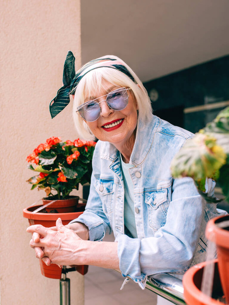 ηλικιωμένη (παλιά) κομψή γυναίκα με γκρίζα μαλλιά και σε μπλε γυαλιά και denim στέκεται στο μπαλκόνι σε εξωτερικούς χώρους. Διακοπές, παρτυ, σπίτι, στύλ, ταξίδια, αντι-ηλικία, διασκεδαστική έννοια - Φωτογραφία, εικόνα