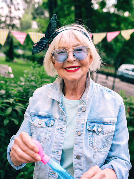senior (old) stylish woman with gray hair and in blue glasses and jeans jacket blowing bubbles outdoors. Holidays, party, anti age, fun concept - Photo, Image