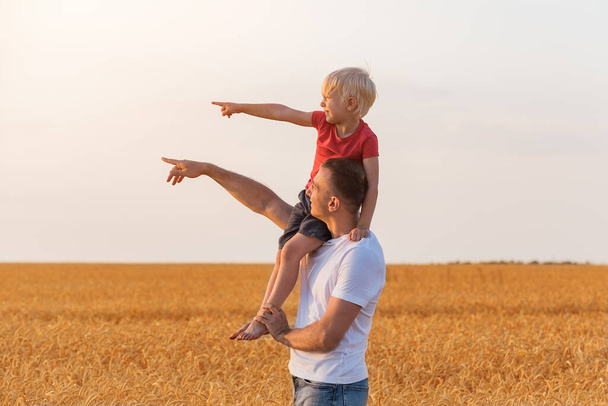 Father and son pointing into the distance. Child sits on his fathers shoulder on background of yellow field. - Foto, afbeelding