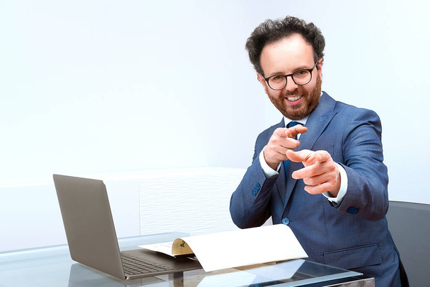 Usmívající se evropský podnikatel v obleku, ukazující prsty před kamerou na interiéru kanceláře se skleněným stolem, notebookem, dokumentem. Koncepce rozvoje podnikání a růstu - Fotografie, Obrázek