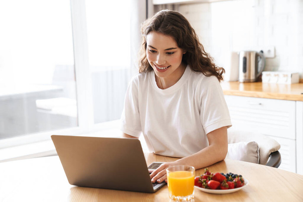 Photo of cheerful beautiful woman using laptop while having breakfast in modern kitchen - Фото, зображення