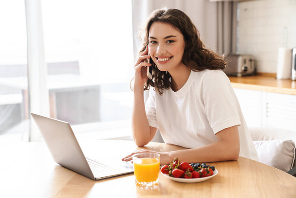 Photo of smiling beautiful woman using laptop and talking on cellphone while having breakfast at kitchen - Foto, imagen