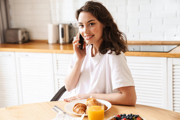Photo of smiling beautiful woman talking on mobile phone while having breakfast at kitchen - Foto, Bild