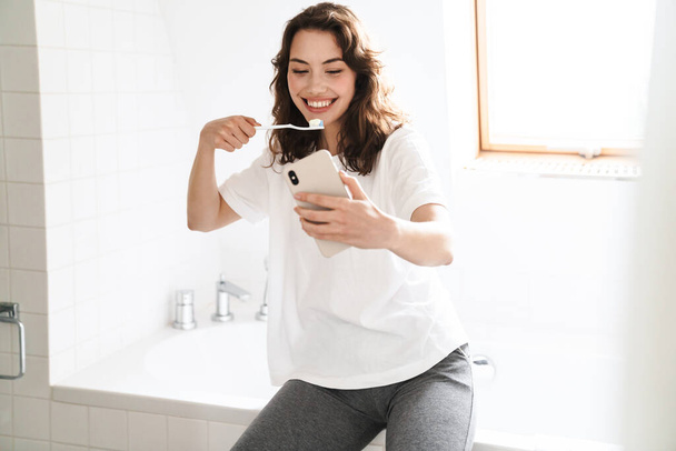 Photo of cheerful beautiful woman smiling and cleaning teethes while using mobile phone at bathroom - Фото, зображення