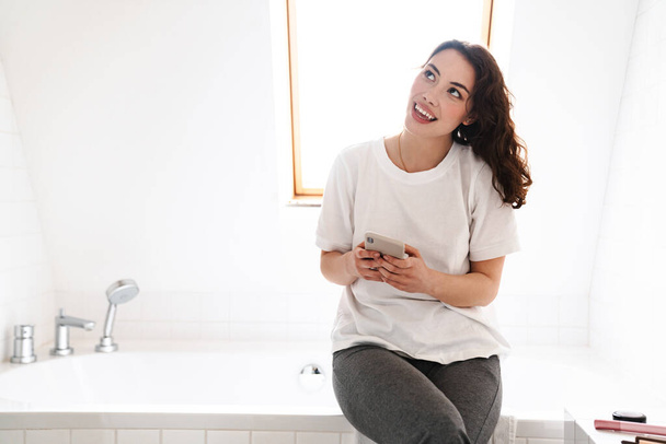Photo of cheerful beautiful woman smiling and using mobile phone while sitting in bathroom - Foto, Imagem