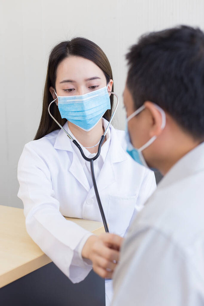 Aziatische man patiënt worden gecontroleerd zijn gezondheid, terwijl een vrouwelijke arts gebruik maken van een stethoscoop om hartslag van hem te horen in Coronavirus pandemie Door het dragen van een chirurgisch masker te allen tijde. Bescherming tegen het coronavirus. - Foto, afbeelding