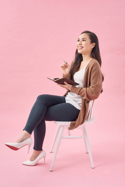 Smiling lovely girl writing in a diary while sitting on a chair isolated over pink wall - Foto, Imagem