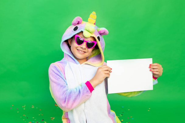 a little girl on a green isolated background in a bright suit holding a white sheet of paper. Space for text. The concept of a holiday, Birthday and sales - Fotó, kép