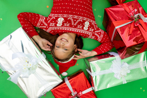mała dziewczynka leży na zielonym, odizolowanym tle w czerwonej kurtce i kapeluszu Mikołaja wśród stosu prezentów. Miejsce na tekst. Koncepcja nowego roku i Bożego Narodzenia - Zdjęcie, obraz