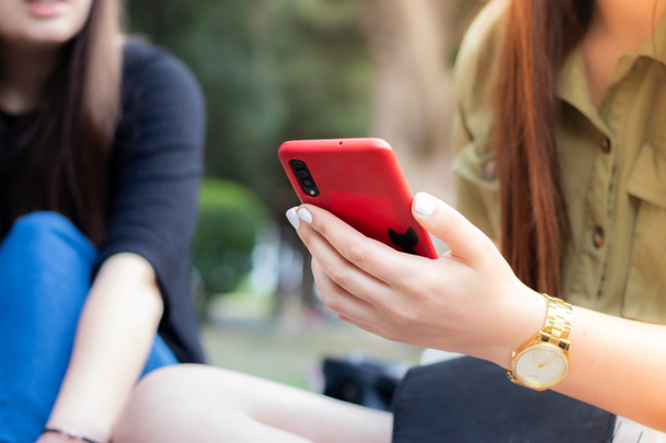 Közelkép a lány kezét fehér körmök kezében egy piros okostelefon egy parkban. A telefonjával tölti az idejét, míg a barátaival kint piknikezik. - Fotó, kép