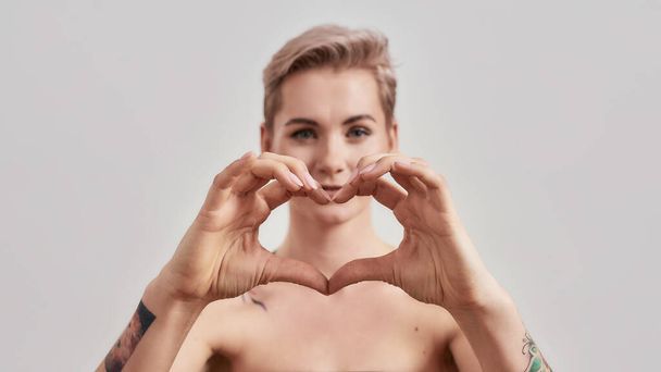 Heart you. Close up portrait of a young attractive half naked tattooed woman with perfect skin looking at camera, showing heart with hands isolated over light background - Foto, Imagem