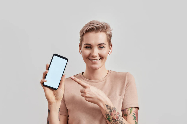 Portrait of tattooed woman with pierced nose and short hair in wireless earbuds or earphones smiling at camera, pointing at smartphone with blank screen isolated over light background - Fotó, kép