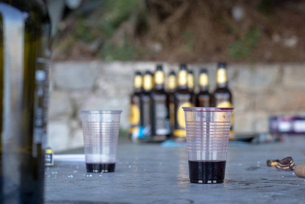 空のワイン2杯。背景にはブルービールボトル。公園内の公共飲料のゴミやゴミの遺跡。公の場で飲む若者と酒の概念 - 写真・画像