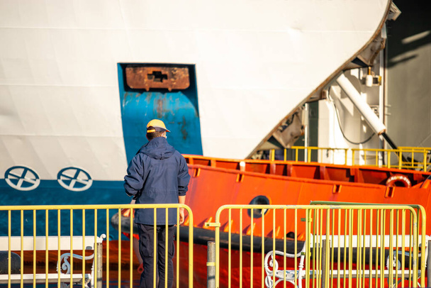 Za žlutým plotem, zpovzdálí spatřen, stojí převozní lodní dělník, v dálce několik obrovských lodí. Část bílého trajektu a oranžového vlečného člunu - Fotografie, Obrázek