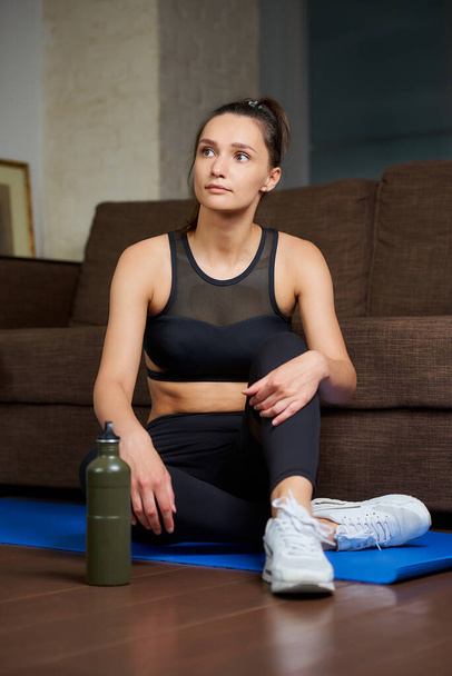 Een sportief meisje in een zwart trainingspak rust tussen de trainingssets op de blauwe yogamat bij een groene aluminium fles water en de bank thuis. - Foto, afbeelding