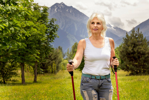 Λεπτή ηλικιωμένη γυναίκα με γκρίζα μαλλιά περπατά στον κήπο με μπαστούνια για το περπάτημα στο φόντο των βουνών σε μια ηλιόλουστη μέρα - Φωτογραφία, εικόνα