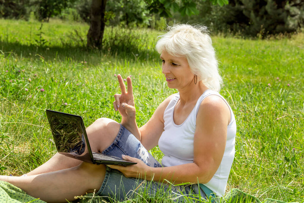 Струнка літня жінка з сірим волоссям сидить на сонячному газоні в парку і спілкується через ноутбук
 - Фото, зображення