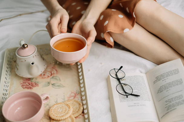 A woman holds a cup of hot tea on the bed at home and reads a book, home leisure in coziness and comfort. High quality photo - Фото, зображення