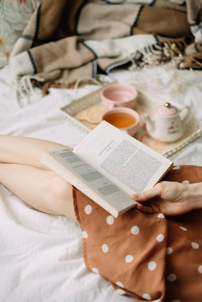 A woman reads a book at home in bed and drinks tea, home leisure in coziness and comfort. High quality photo - 写真・画像