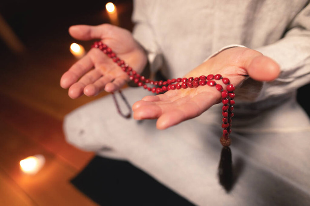 close-up man in clothes for practice and meditation sits in a lotus pose and holds red rosary to concentrate attention in a wooden room with dim light - Foto, imagen