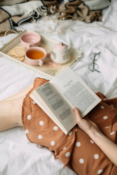 A woman reads a book at home in bed and drinks tea, home leisure in coziness and comfort. High quality photo - Foto, afbeelding