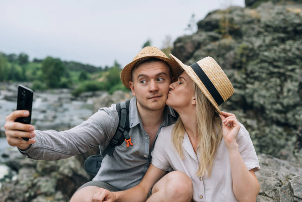 Счастливая молодая пара путешественников в повседневной одежде делает селфи на мобильном телефоне на фоне горной реки. Местный туризм, концепция поездки на выходные - Фото, изображение