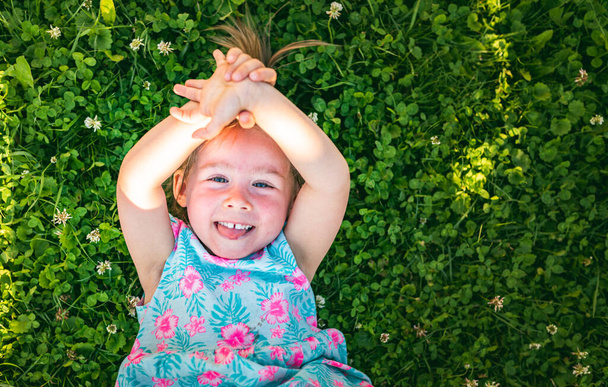 Mooi klein meisje liggend op gras lachend en omhoog kijkend. 2-3 jaar oud kind in de natuur op zonnige zomerdag - Foto, afbeelding