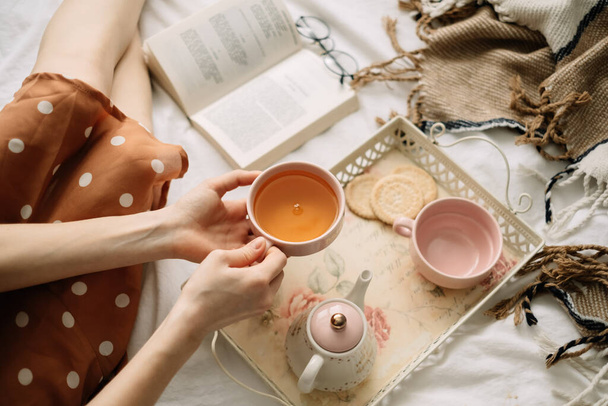 A woman holds a cup of hot tea on the bed at home and reads a book, home leisure in coziness and comfort. High quality photo - Photo, image