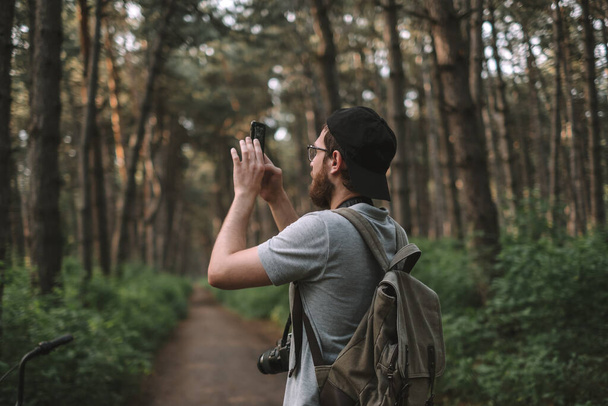 A young male tourist takes pictures on a smartphone camera in a forest. Tourism, active lifestyle. High quality photo - Foto, Imagen