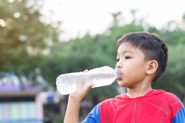 Muotokuva lapsesta 5-6 vuotta vanha. Onnellinen aasialainen lapsi opiskelija poika juo vettä muovipullosta. Lopetettuaan harjoittelun. Terveellinen lapsi ja ruokaa. Kesäkaudella. Vihreä tausta. - Valokuva, kuva