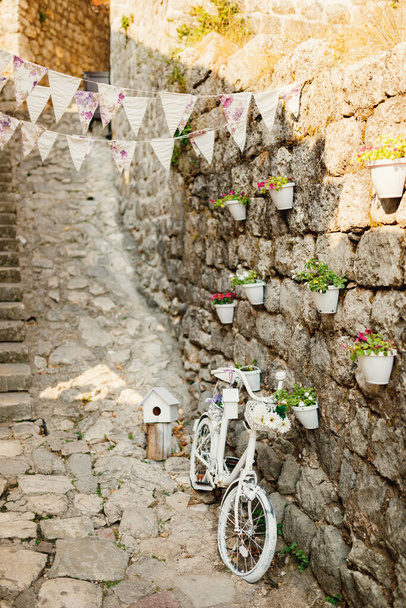 Белый декоративный велосипед у стены с горшками для цветов и флагов. - Фото, изображение