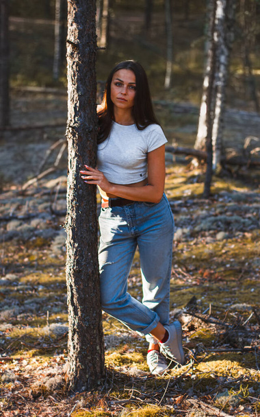 Молодая модная красотка в маминых джинсах и белом топе прислоняется и держит руку на дереве в летнем сосновом лесу - Фото, изображение