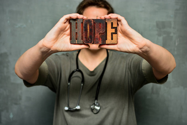 Een medisch medewerker met een stethoscoop heeft het woord "hoop" in zijn hand, gemaakt van houten typografische letters. Gezondheidszorgpersoneel, dokter, verpleegkundige, hoogwaardig concept van medische zorg. - Foto, afbeelding