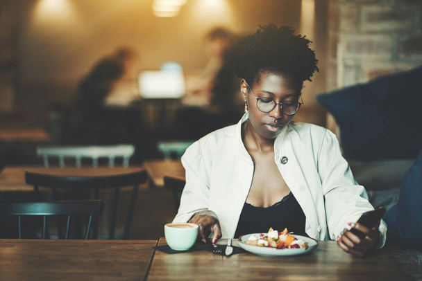 Портрет с копировальным пространством слева от молодой изящной черной женщины в элегантных очках и белой траншеи, обедающей в уютном ресторанном укромном уголке и пользующейся смартфоном - Фото, изображение