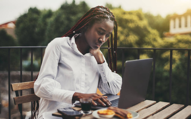 A ravishing young black woman freelancer is working remotely with her project using a laptop while sitting at the table with a cup of coffee and delicious bakery on the balcony of a street restaurant - Fotoğraf, Görsel