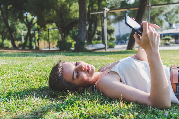 beautiful young caucasian woman laying on the grass of an urban park and using a telephone - Foto, Imagem