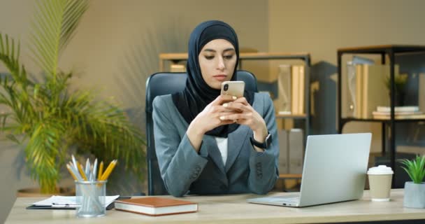 Arabian beautiful young woman in hijab sitting at table in cozy office and tapping or scrolling on smartphone in front of laptop computer. Muslim businesswoman texting message on mobile phone. - Séquence, vidéo