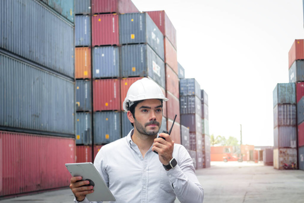 Hombre de negocios guapo con casco de seguridad, usando walkie talkie y mantener la mesa digital durante la inspección en el astillero de contenedores de carga de envío logístico
 - Foto, imagen