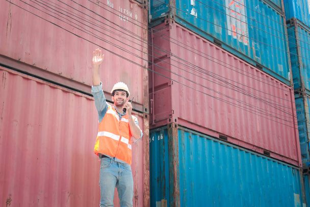 Trabajador capataz guapo que usa equipo de seguridad, hablando en el walkie-talkie durante la inspección en el astillero de contenedores de carga de envío logístico
 - Foto, imagen