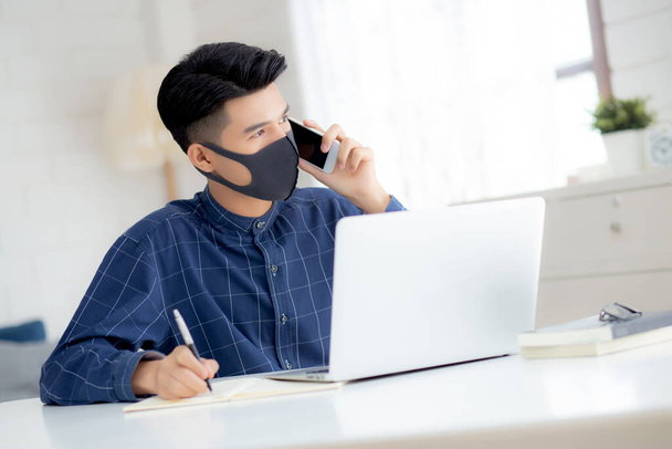 Młody azjatycki biznesmen w masce twarzy mówi smartfon i pracuje na laptopie do ochrony covid-19, kwarantanny biznesmen pracy z domu i pisania notebooka, odległość społeczna i pobyt w domu. - Zdjęcie, obraz