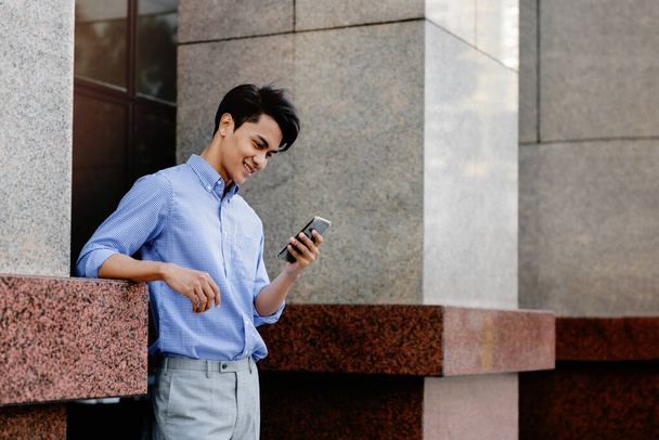 Usmívající se mladý asijský podnikatel pomocí mobilního telefonu ve městě - Fotografie, Obrázek