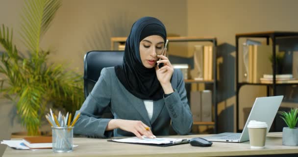 Arabian woman in hijab sitting at desk in office, talking on cellphone and working on laptop. Female muslim speaking on phone and noting at documents with charts and graphics. Arab businesswoman. - Filmagem, Vídeo