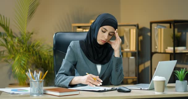 Tired Arabian woman in hijab sitting at desk in office, thinking and working on laptop. Female muslim manager typing on computer and noting at documents with charts and graphics. Arab businesswoman. - Filmagem, Vídeo